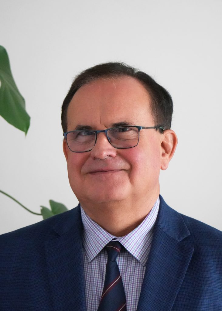 Mężczyzna w średnim wieku, w okularach, w koszuli z krawatem i w marynarce, widoczny do ramion. W tle widocznych częściowo kilka zielonych liści