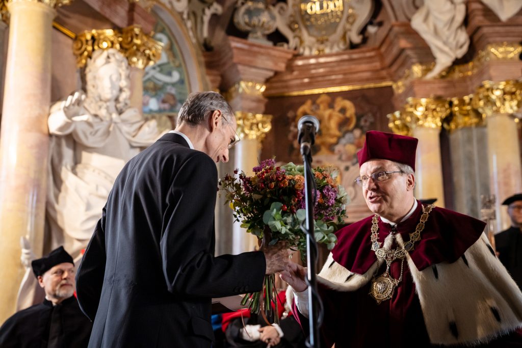 Dwóch mężczyzn w średnim wieku, jeden po lewej stronie ubrany w garnitur, odwrócony bokiem, w dłoni trzyma bukiet czerwonych róż, drugi po prawej stronie w stroju rektorskim w kolorach białym i bordowym, w bordowej czapce.