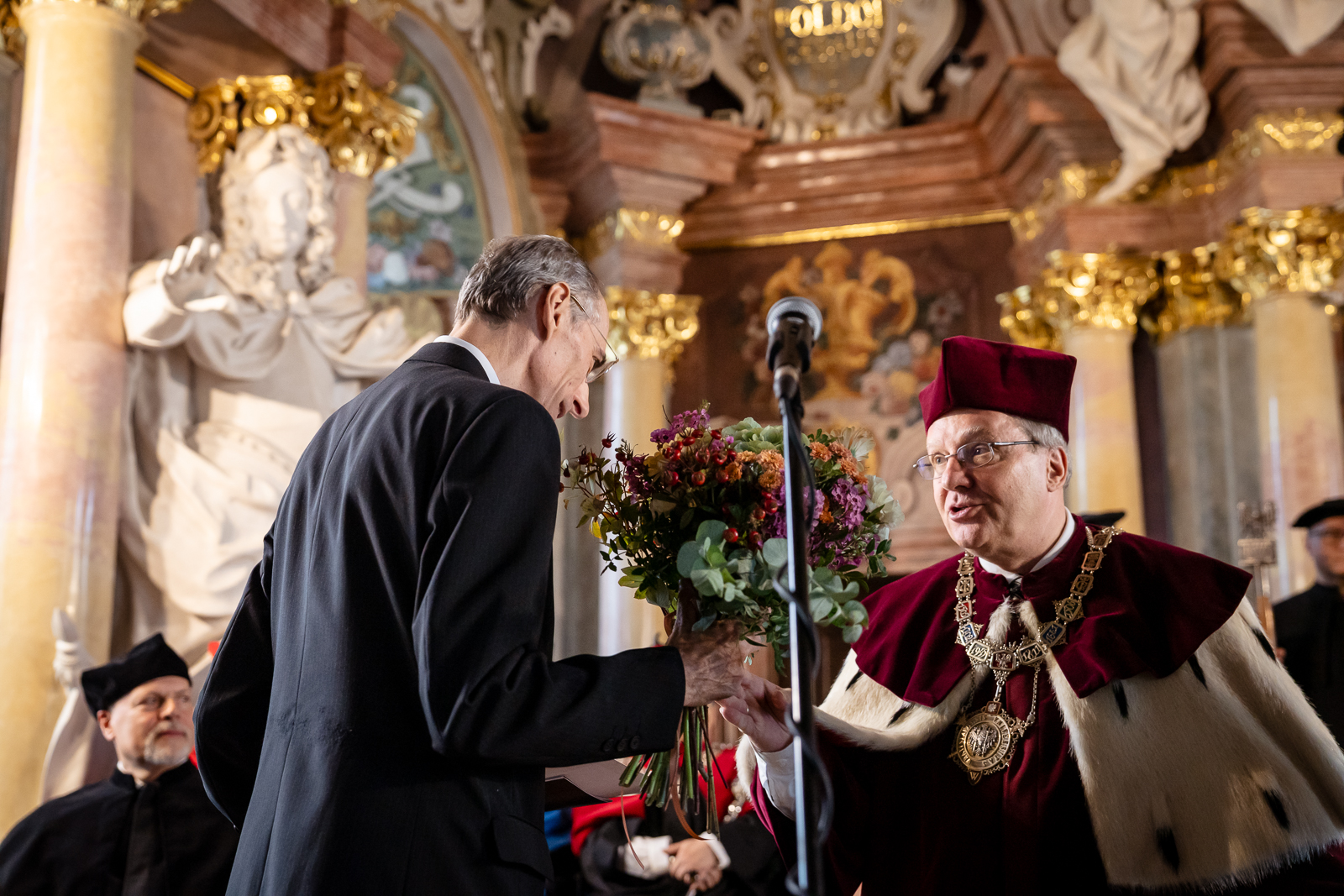 Dwóch mężczyzn w średnim wieku, jeden po lewej stronie ubrany w garnitur, odwrócony bokiem, w dłoni trzyma bukiet kwiatów, drugi po prawej stronie w stroju rektorskim w kolorach białym i bordowym, w bordowej czapce.