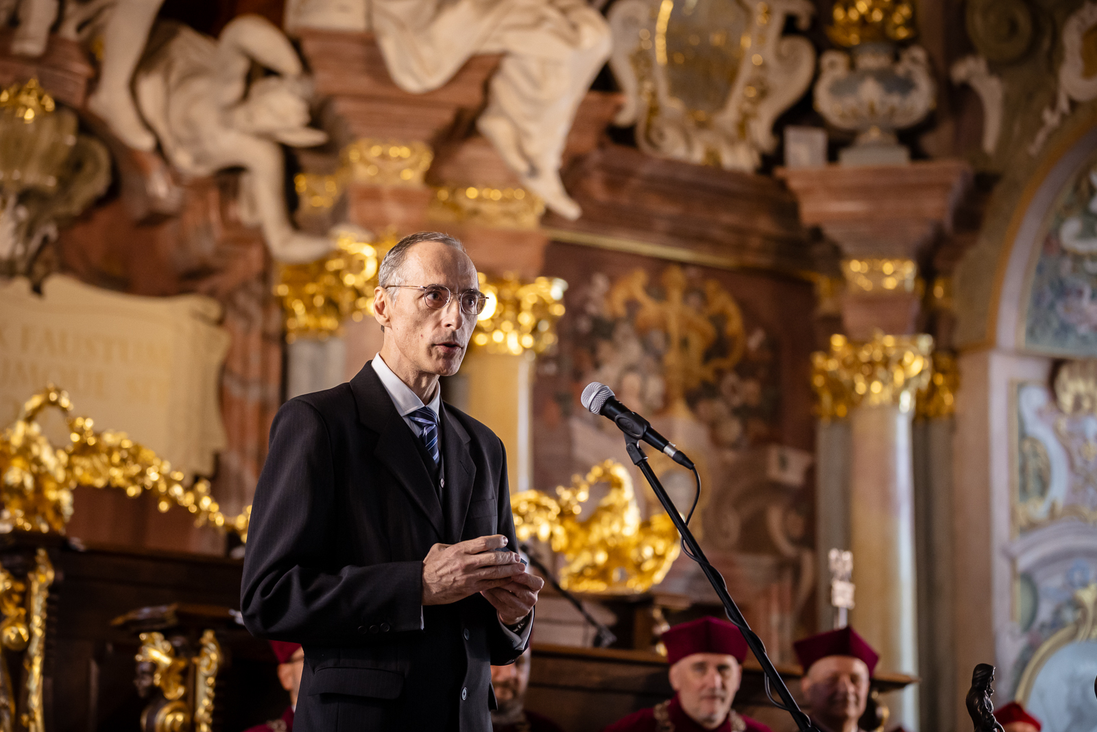 Po lewej stronie mężczyzna w średnim wieku, w okularach, widoczny do pasa, ubrany w marynarkę, po prawej stronie statyw z mikrofonem, w tle ściana z kolumnami z rzeźbami i ozdobami w kolorze złotym.